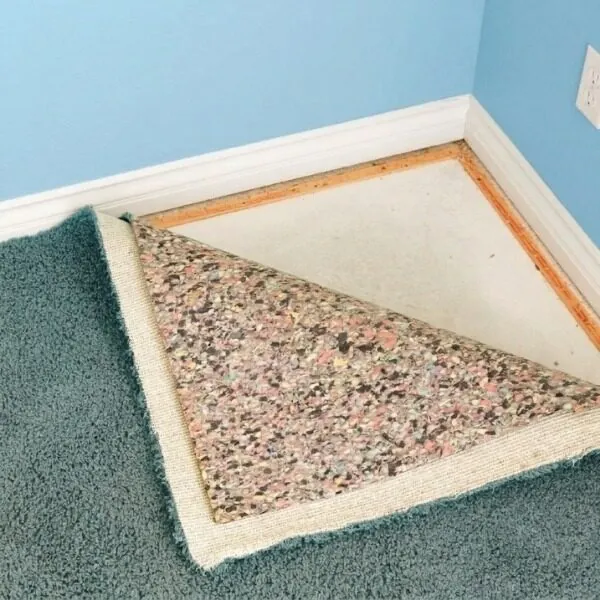 A room corner showing a carpet underlay installation process, with the teal carpet pulled back to display the layered speckled foam underlayment and smooth edge tack strips.