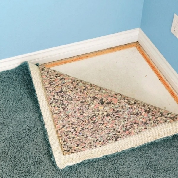 A room corner showing a carpet underlay installation process, with the teal carpet pulled back to display the layered speckled foam underlayment and smooth edge tack strips.